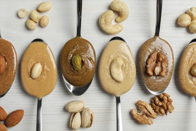 Tasty nut butters in spoons and raw nuts on white wooden table, flat lay