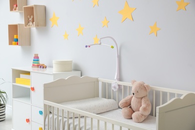 Crib with toy bear and mobile in stylish baby room interior
