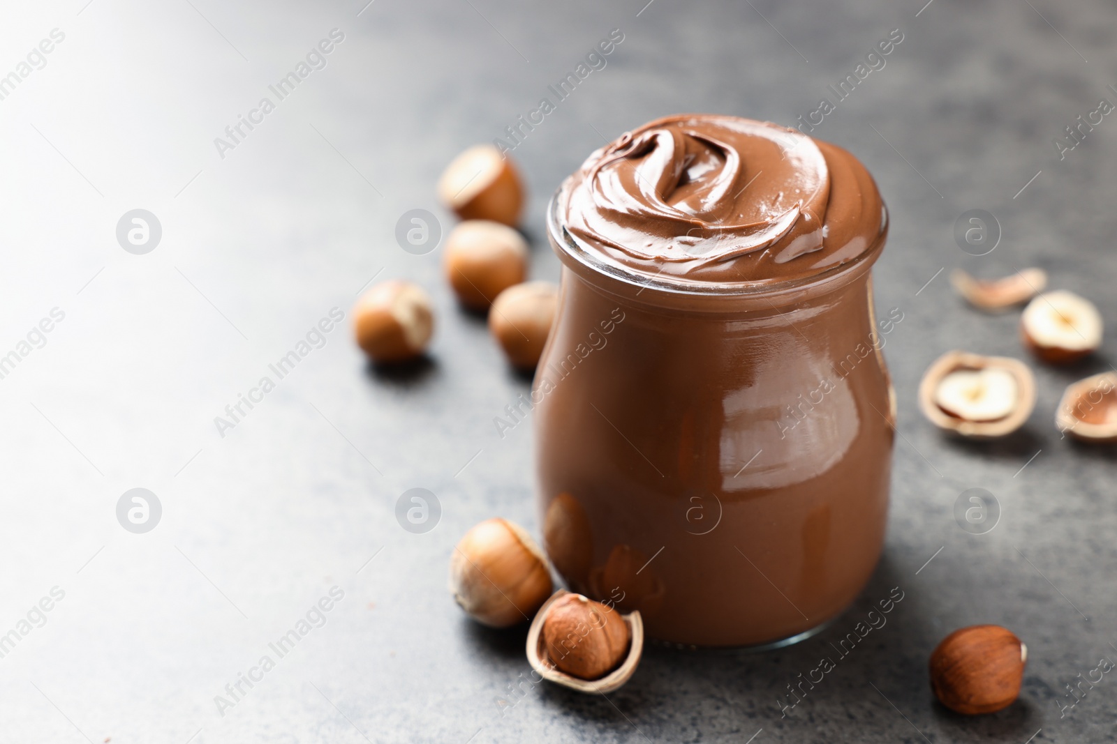 Photo of Glass jar with tasty chocolate hazelnut spread and nuts on grey table. Space for text