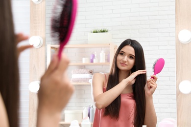 Beautiful young woman with hair brush looking into mirror in bathroom