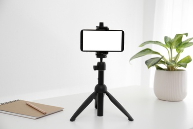 Smartphone with blank screen fixed to tripod on white table indoors. Mockup for design