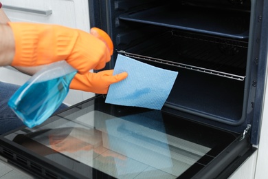 Young man cleaning oven with rag and detergent in kitchen, closeup