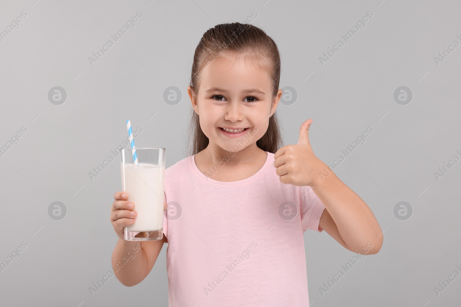 Photo of Cute girl with glass of fresh milk showing thumb up on light grey background