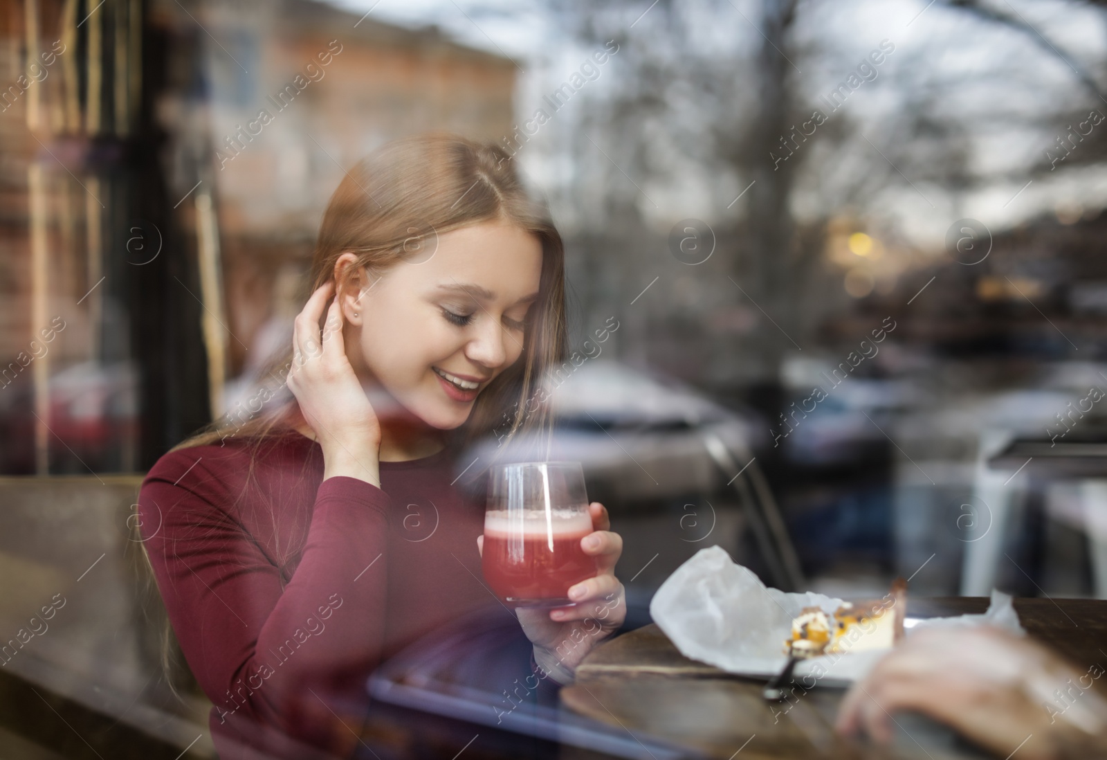 Photo of Pretty young woman with cocktail and cake at table in cafe, view from outdoors through window