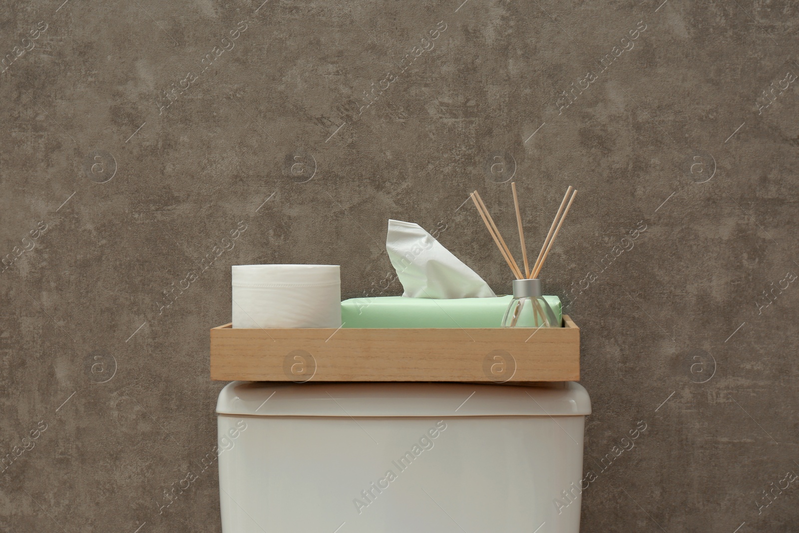 Photo of Tray with roll of paper, napkins and reed air freshener on toilet tank near grey wall. Bathroom interior