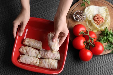 Woman putting uncooked stuffed cabbage roll into baking dish at black table, top view