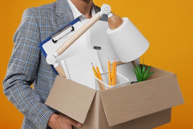Photo of Unemployed young man with box of personal office belongings on orange background, closeup