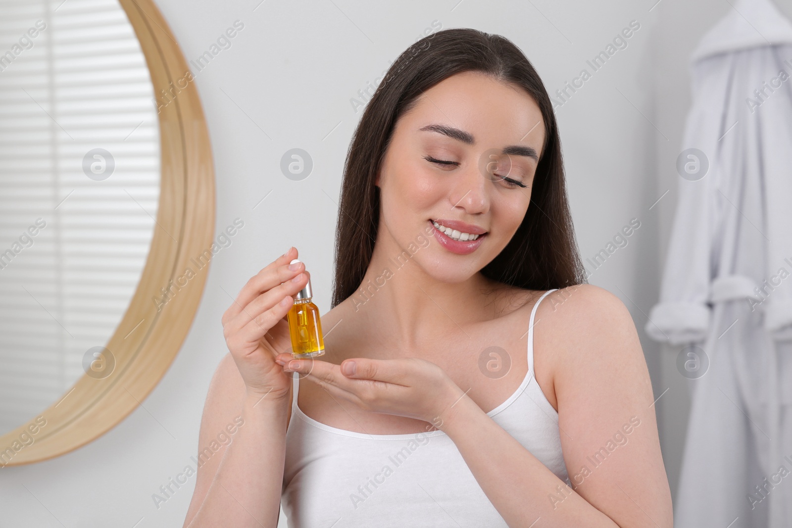 Photo of Happy young woman with bottle of essential oil in bathroom