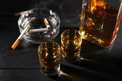 Alcohol addiction. Whiskey in glasses, smoldering cigarette and ashtray on black wooden table