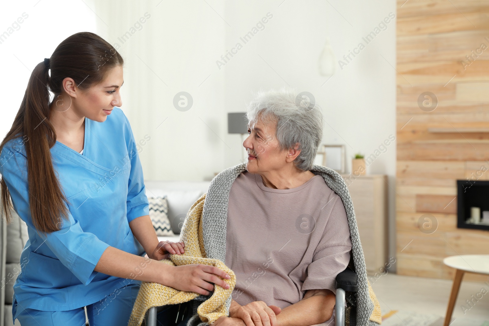 Photo of Nurse covering elderly woman in wheelchair with blanket indoors. Assisting senior people