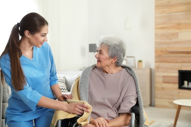 Nurse covering elderly woman in wheelchair with blanket indoors. Assisting senior people