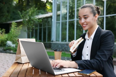 Photo of Happy businesswoman with sandwich using laptop while having lunch at wooden table outdoors