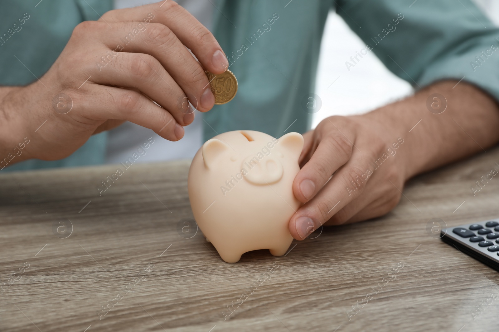 Photo of Man putting coin into piggy bank at wooden table, closeup