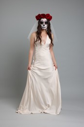 Young woman in scary bride costume with sugar skull makeup and flower crown on light grey background. Halloween celebration