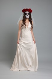 Photo of Young woman in scary bride costume with sugar skull makeup and flower crown on light grey background. Halloween celebration