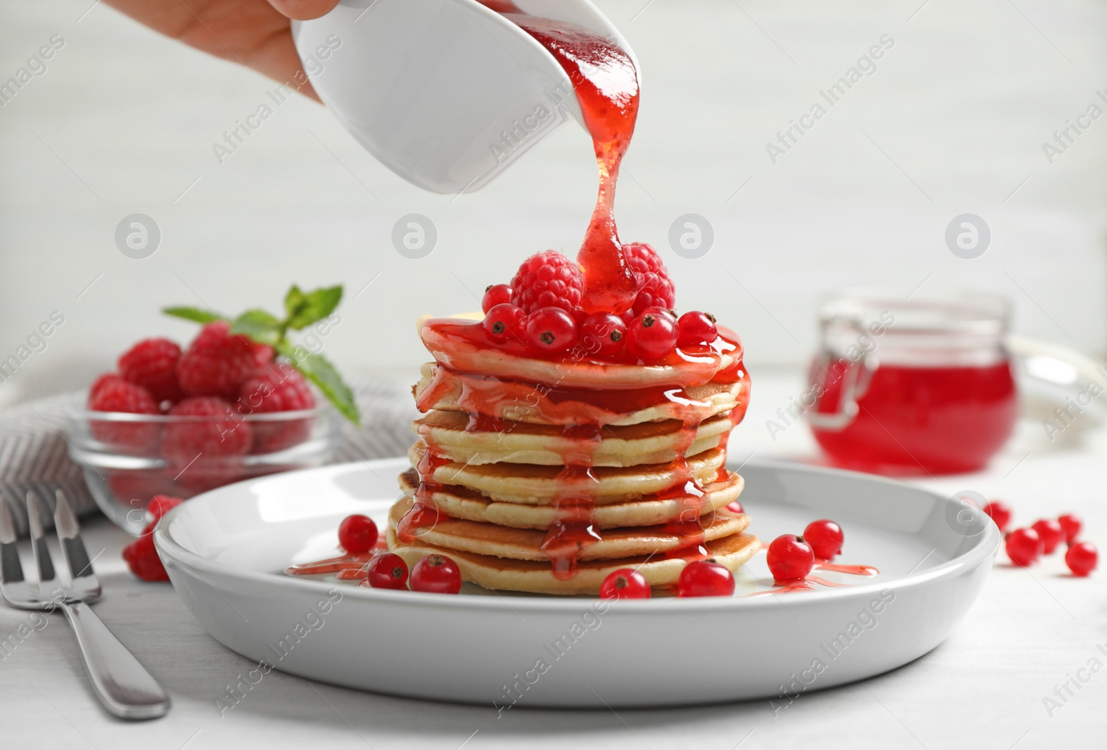 Photo of Woman pouring syrup onto fresh pancakes with berries on white table, closeup