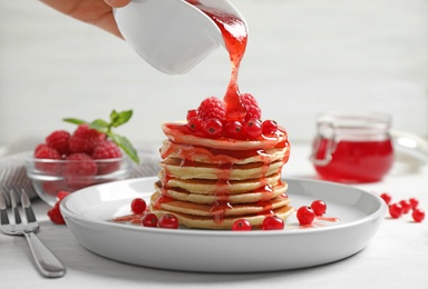 Woman pouring syrup onto fresh pancakes with berries on white table, closeup