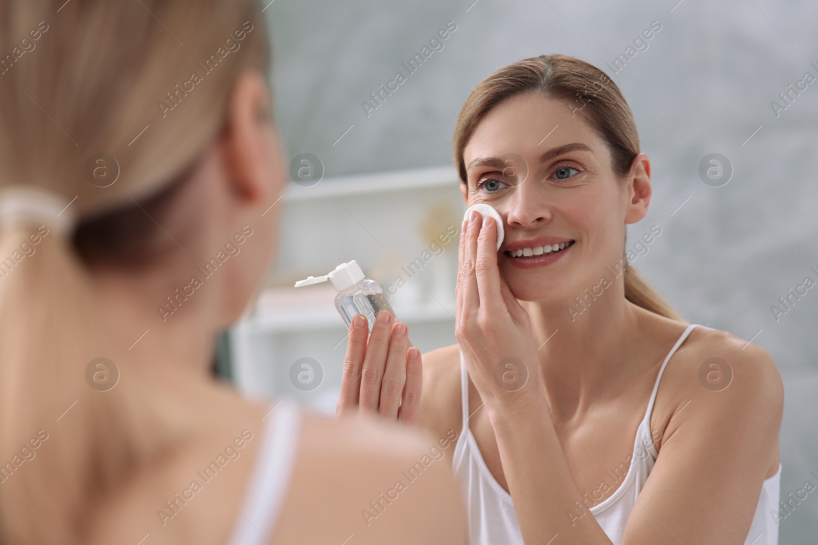 Photo of Beautiful woman removing makeup with cotton pad near mirror indoors