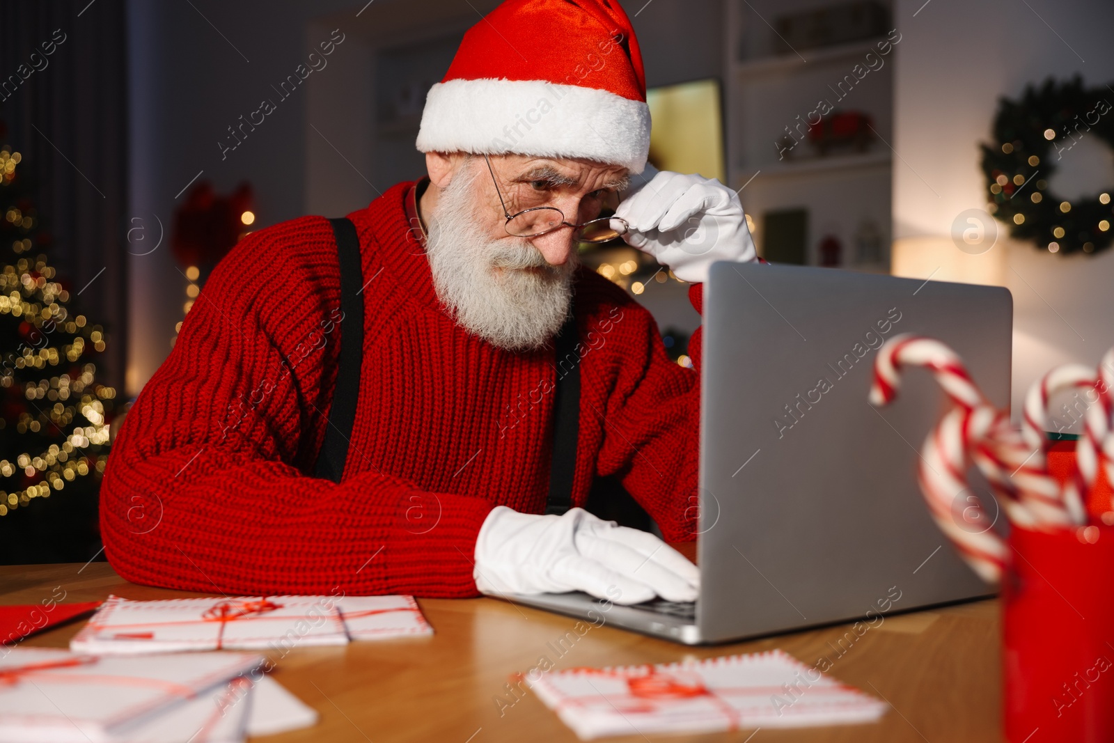 Photo of Santa Claus using laptop at his workplace in room decorated for Christmas