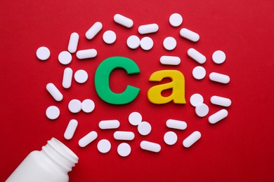 Photo of Pills, open bottle and calcium symbol made of colorful letters on red background, flat lay