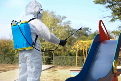 Person in hazmat suit with disinfectant sprayer near slide at children's playground. Surface treatment during coronavirus pandemic