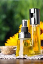 Spray bottles with cooking oil near sunflower seeds and flowers on wooden table against blurred green background