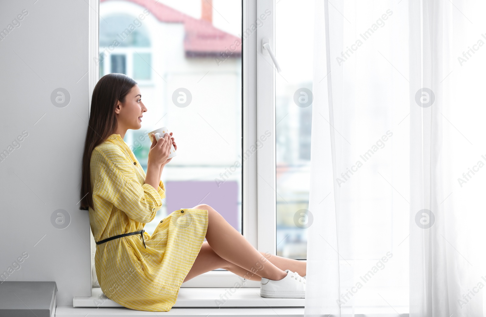 Photo of Young woman with cup on window sill indoors