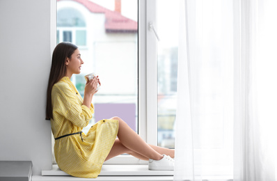 Photo of Young woman with cup on window sill indoors