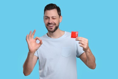 Happy man with condom showing ok gesture on light blue background