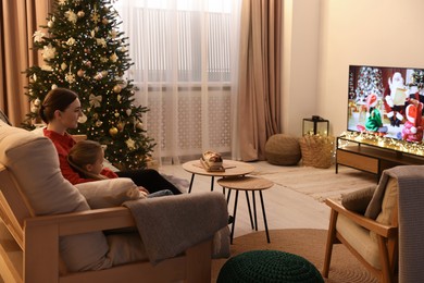 Photo of Mother with her daughter watching TV in cosy room. Christmas atmosphere