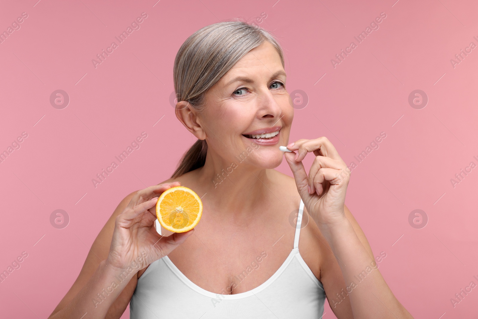 Photo of Beautiful woman with half of orange taking vitamin capsule on pink background