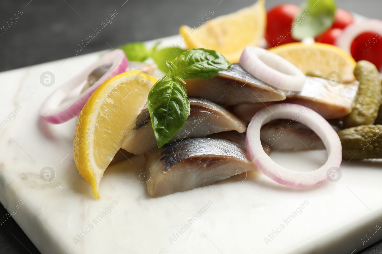 Photo of Serving board with sliced salted herring fillet, basil, onion rings, pickles and lemon on table, closeup