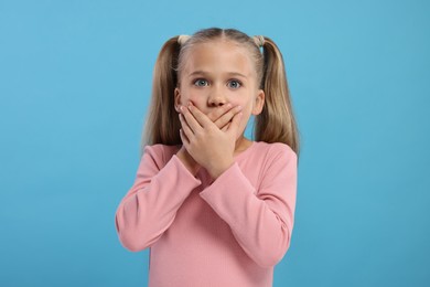 Embarrassed little girl covering her mouth with hands on light blue background