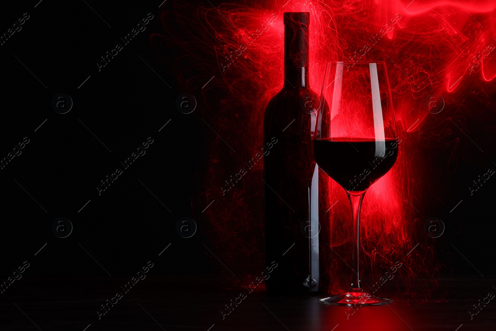 Photo of Tasty wine in glass and bottle in red lights on black background, space for text