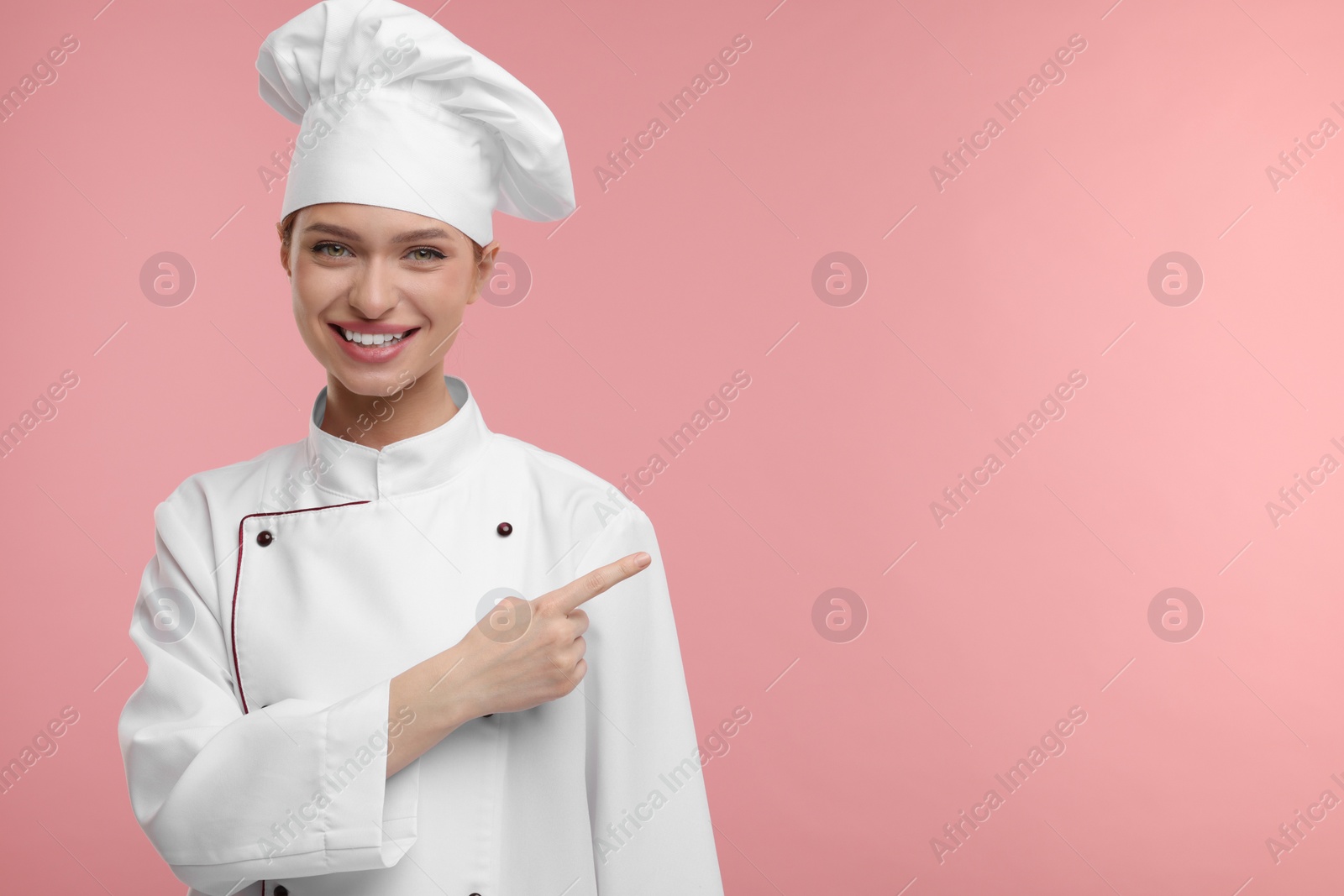 Photo of Happy chef in uniform pointing at something on pink background, space for text