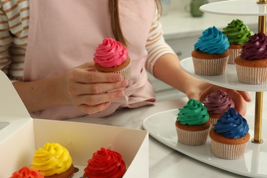 Woman putting delicious colorful cupcakes on dessert stand at white table indoors, closeup