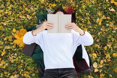 Photo of Woman reading book on grass with dry leaves, top view. Autumn season