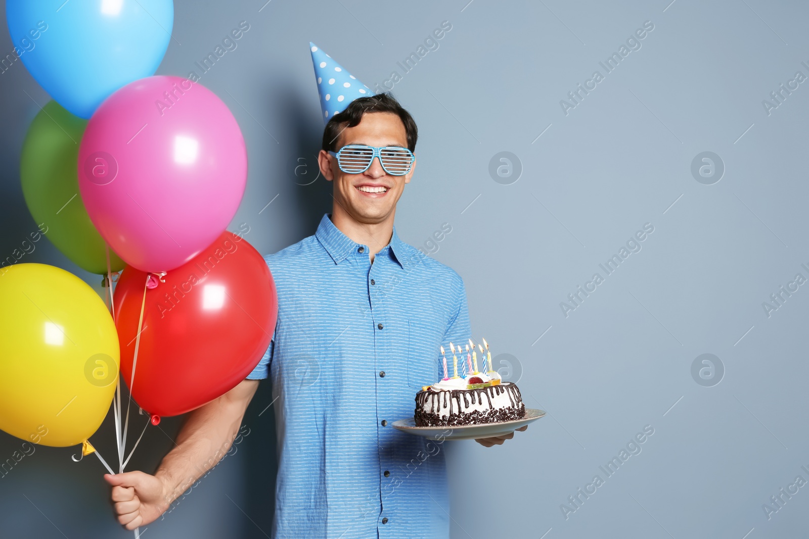 Photo of Young man with birthday cake and bright balloons on color background