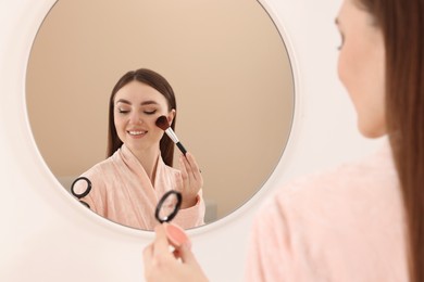 Beautiful young woman applying blusher with brush near mirror indoors