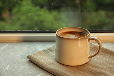 Photo of Cup of hot drink on window sill against glass with rain drops, space for text