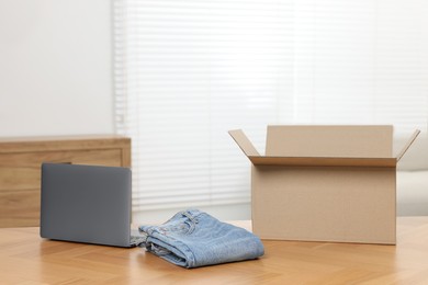 Stylish jeans, modern laptop and cardboard box on wooden table indoors. Online shopping