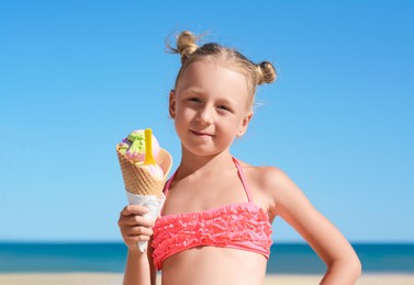 Photo of Adorable little girl in swimsuit with delicious ice cream at beach on sunny summer day