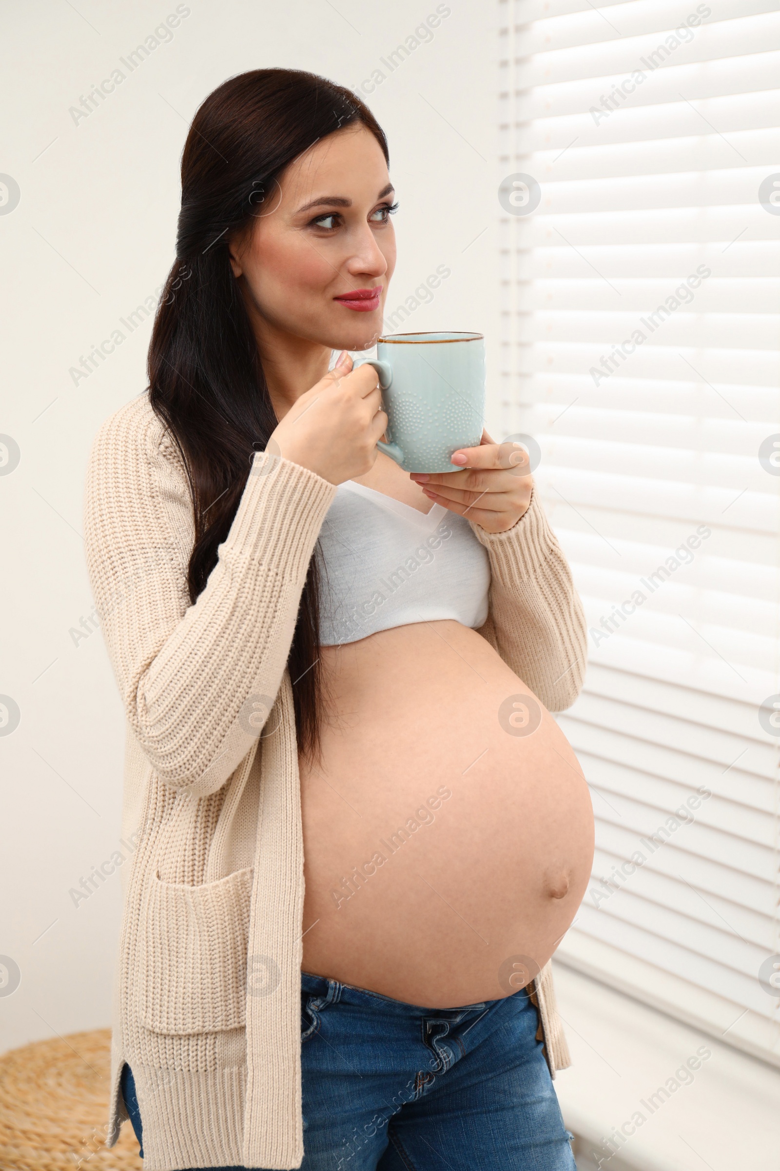 Photo of Beautiful pregnant woman with cup of hot drink near window at home