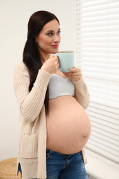 Photo of Beautiful pregnant woman with cup of hot drink near window at home