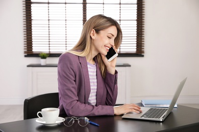 Image of Young woman talking on phone while using laptop in office
