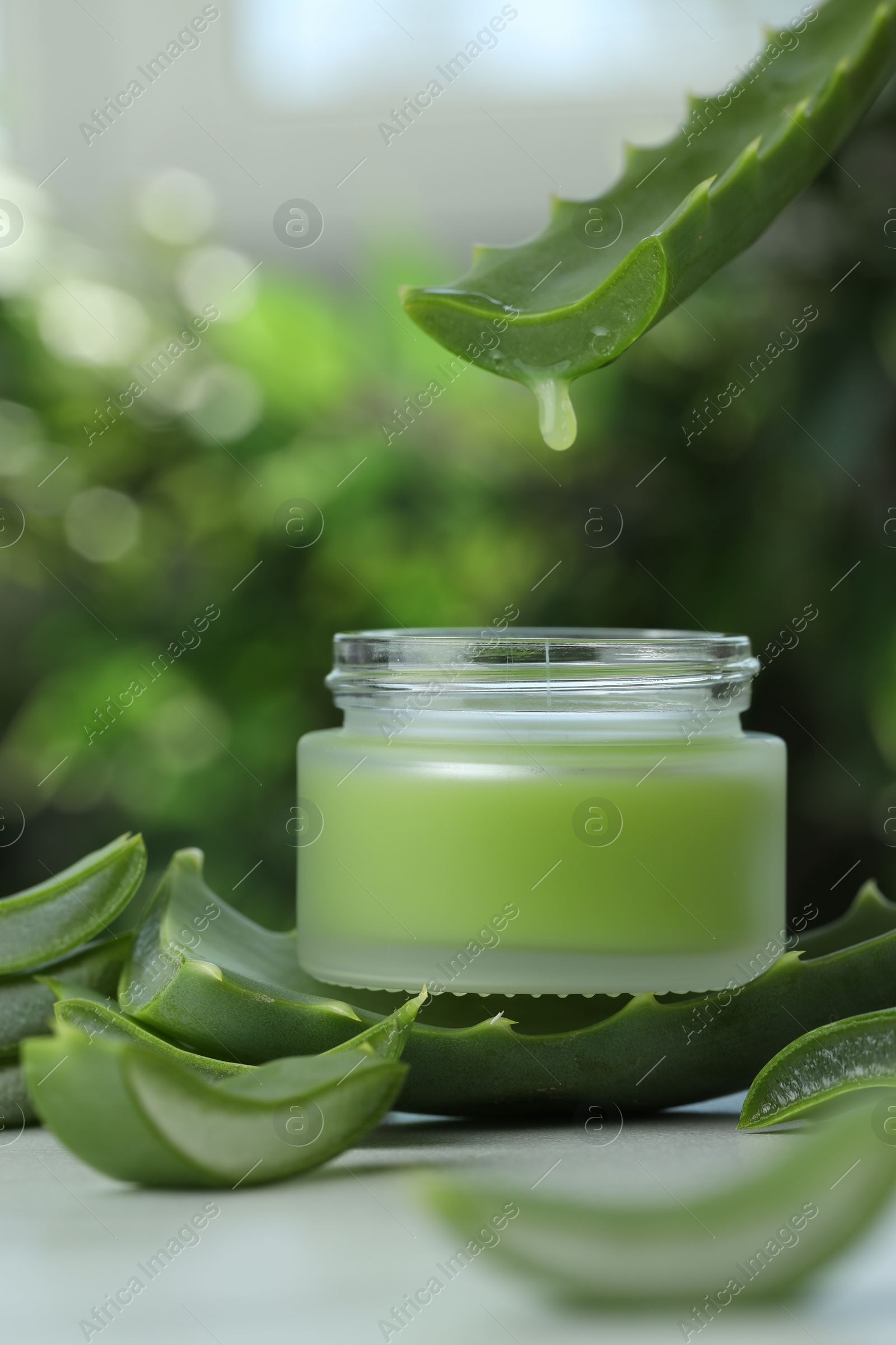 Photo of Aloe vera juice dripping from leaf into jar with cream on white table against blurred background