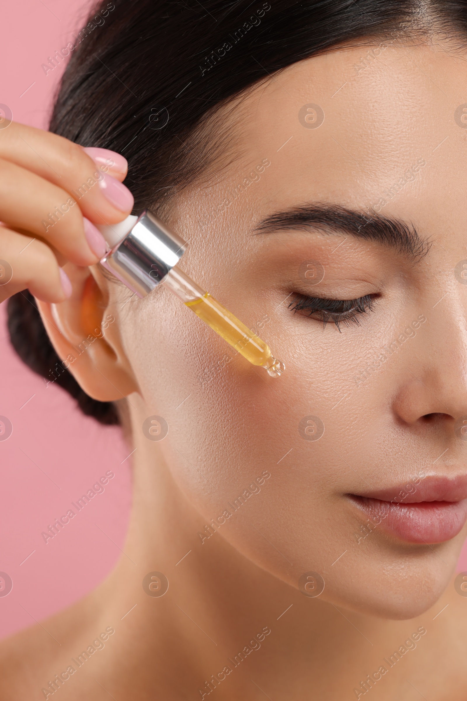 Photo of Beautiful young woman applying serum onto her face on pink background, closeup