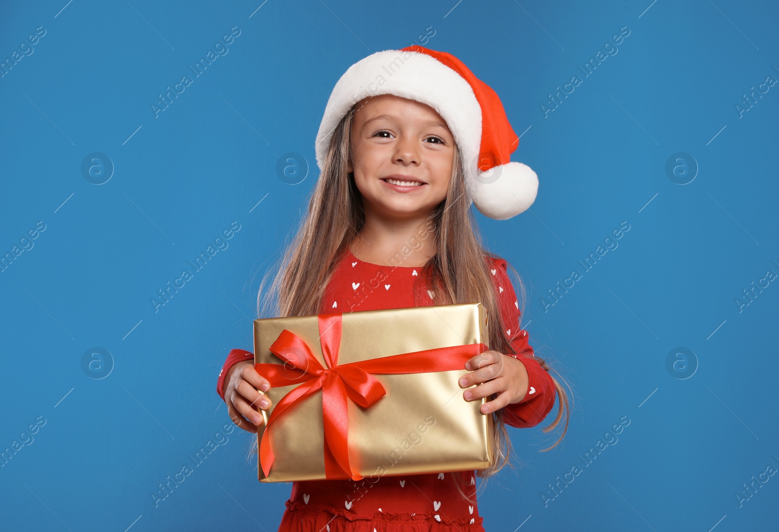 Photo of Happy little child in Santa hat with gift box on blue background. Christmas celebration