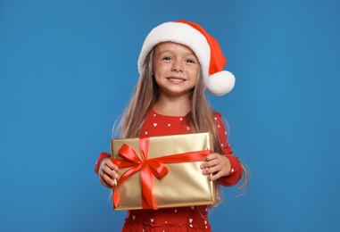 Photo of Happy little child in Santa hat with gift box on blue background. Christmas celebration