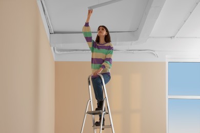 Photo of Young woman installing ceiling lamp on stepladder indoors. Room renovation